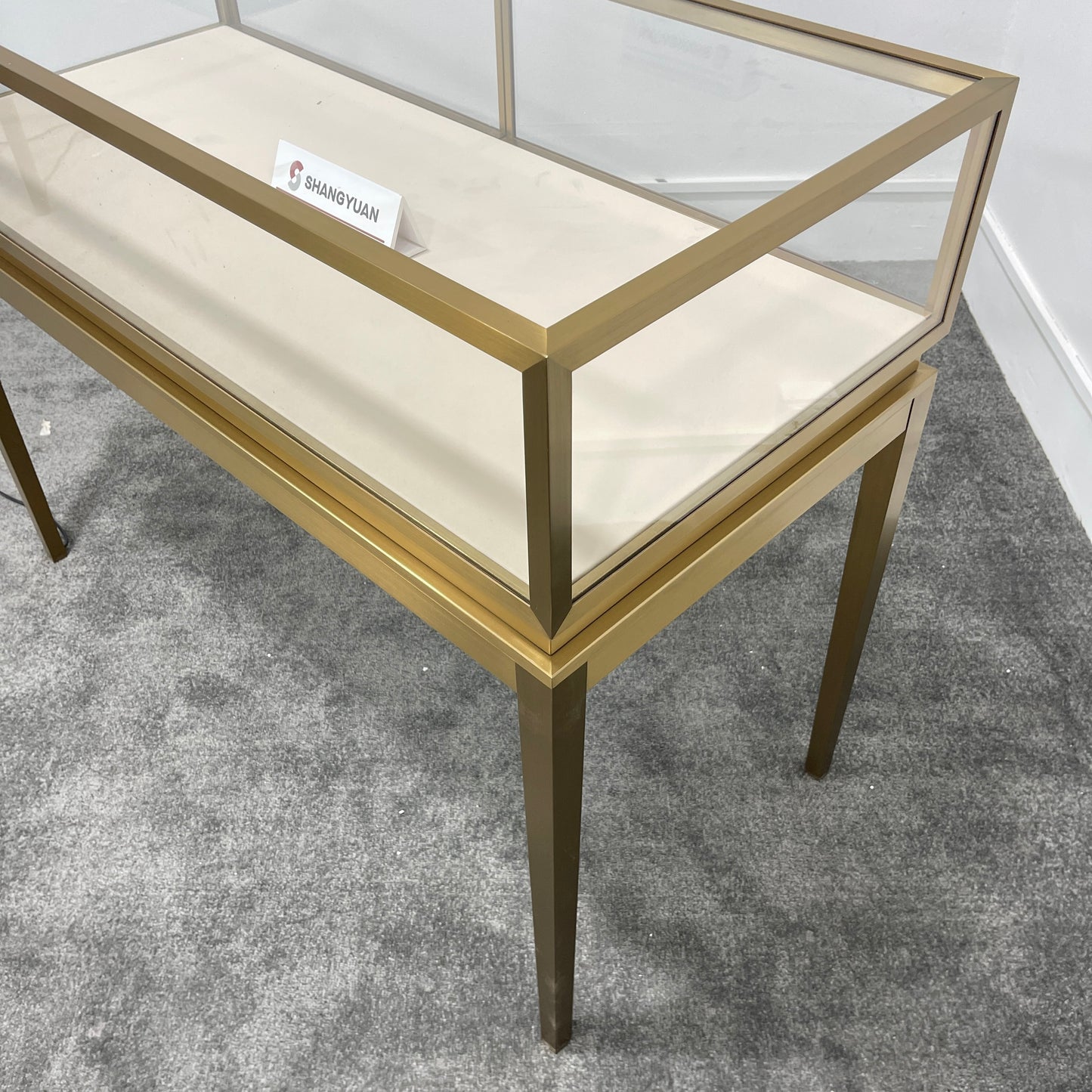 Close-up corner detail of gold brushed stainless steel jewelry display case with tempered glass top and cream velvet interior